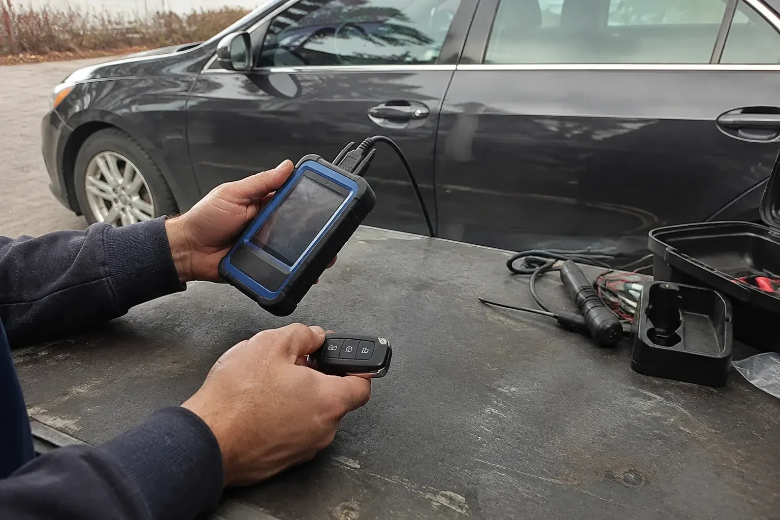 Hands holding a blue-edged electronic device and car-key fob at a worktable beside a parked car. — Transponder Key Programming, Haldimand County