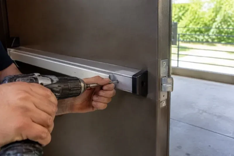 Hands using a power driver at the end of a horizontal push bar on an open metal door. — Panic & Exit Hardware, Haldimand County